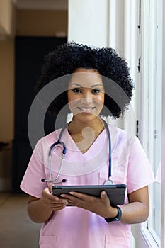 Biracial nurse holding tablet, wearing pink scrubs, standing by window at home