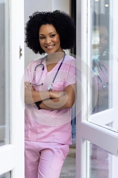 Biracial nurse standing by door at home, arms crossed, wearing pink scrubs