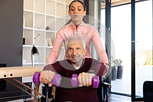 Biracial physiotherapist assisting caucasian senior man sitting on wheelchair in lifting dumbbells