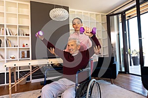 Biracial physiotherapist assisting senior caucasian man sitting on wheelchair in lifting dumbbells