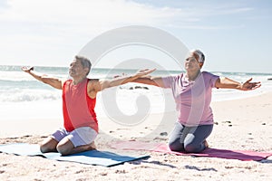 Biracial senior couple kneeling while practicing yoga with arms outstretched on mats at sunny beach