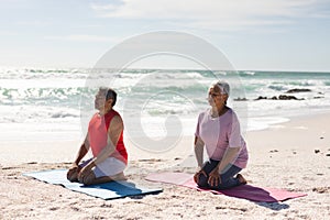 Biracial senior couple meditating while kneeling on yoga mats at beach against sky during sunny day