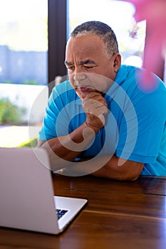 Biracial senior man with hand on chin looking at laptop on table while sitting in nursing home