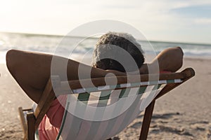 Biracial senior man relaxing with hands behind head sitting on folding chair at beach during sunset