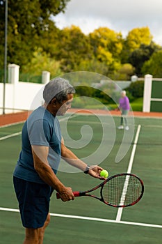 Biracial senior man serving tennis ball to senior wife while playing tennis in court against trees