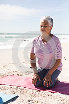 Biracial senior woman with eyes closed meditating while kneeling on yoga mat at sunny beach