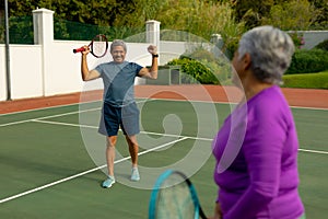 Biracial senior woman looking at cheerful senior man holding racket and gesturing in tennis court