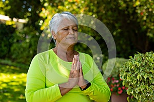 Biracial senior woman with short hair meditating with eyes closed in prayer position at yard