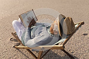 Biracial senior woman wearing hat reading book while relaxing on folding chair at beach