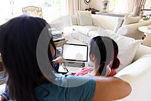 Biracial sisters sitting on couch in living room using laptop, with copy space on screen