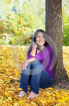 Biracial teen girl leaning against tree, autumn leaves on groun