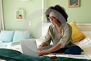 Biracial woman doing paperwork using laptop and smartphone on bed in bedroom