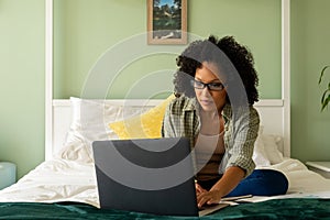 Biracial woman doing paperwork using laptop and smartphone on bed in bedroom