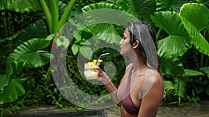 Biracial woman enjoys refreshing cocktail by rich poolside. Relaxed, she sips a tropical drink, feels summer vibe beside