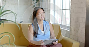 Biracial woman sits comfortably on a yellow sofa, holding a tablet