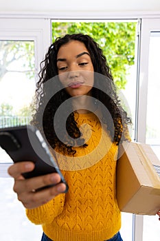 Biracial woman in yellow sweater holding package and using smartphone at home