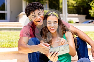 Biracial young couple laughing and taking selfie with mobile phone while sitting on steps in yard