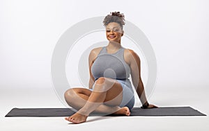 A biracial young female plus size model sits on yoga mat, smiling, copy space
