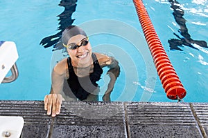 Biracial young female swimmer is hanging onto the pool edge indoors, smiling photo