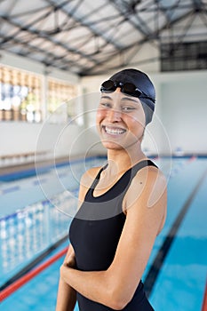 Biracial young female swimmer standing by pool indoors, arms crossed, smiling