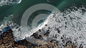 Bird's eye view of ocean waves crashing against rock beach from above. Top view waves break on empty stones in a blue