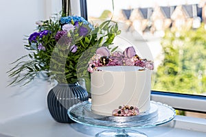 Birthdag cake decorated with pink flowers and a bouguet of flowers in a vase on the windowsill.