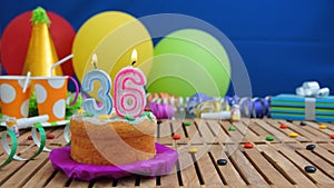 Birthday 36 cake with candles on rustic wooden table with background of colorful balloons, gifts, plastic cups and candies