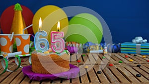 Birthday 65 cake with candles on rustic wooden table with background of colorful balloons, gifts, plastic cups and candies