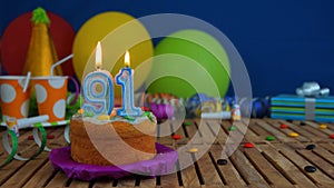 Birthday 91 cake with candles on rustic wooden table with background of colorful balloons, gifts, plastic cups and candies