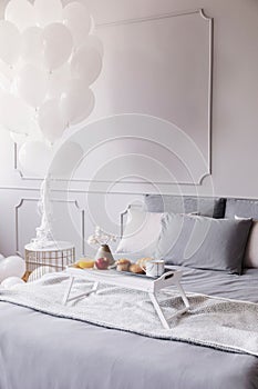 Birthday breakfast on white wooden trey on large bed with grey sheets and blanket, bunch of white balloons above nightstand, copy
