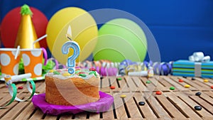 Birthday cake with candle in the shape of a question mark on rustic wooden table with background of colorful balloons
