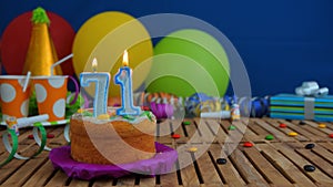 Birthday 71 cake with candles on rustic wooden table with background of colorful balloons, gifts, plastic cups and candies