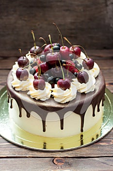 Birthday cake decorated with melted chocolate, whipped cream and fresh cherries. Rustic wooden background.