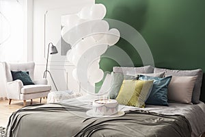Birthday cake on bed with grey duvet and colorful pillows in bright interior with bunch of white balloons and copy space on