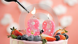 Birthday cake number 89, pink candle on beautiful cake with berries and lighter with fire against background of white