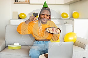 Birthday concept. Joyful black woman having online b-day party, holding pie with burning candle and glass of wine