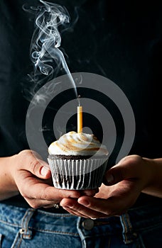 Birthday cupcake with smoking blown out candle in hands of a woman who is wearing black shirt