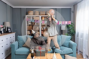 Birthday euphoria. Young couple is sitting at home waiting for birthday party guests where the young woman is jumping on the sofa