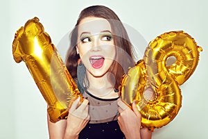 A birthday girl on her 18th birthday with gold number baloons. Excited eighteen girl with green make up and black dress smile.