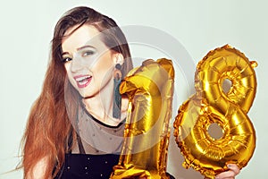 A birthday girl on her 18th birthday with gold number baloons. Excited eighteen girl with green make up and black dress smile.