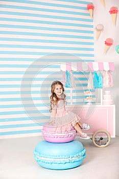 Birthday and happiness concept - happy little girl sitting on a big cake against the background of a candy bar. Decorated room for