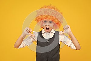 Birthday kids party. Funny kid in curly clown wig isolated on yellow background. Angry teenager girl, upset and unhappy