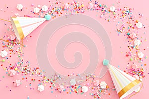 Birthday party background with hats and colorful candies over pastel pink background. Top view