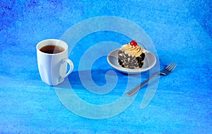 A biscuit cake with chocolate chips and cherries on a cream hat on a white ceramic plate and a cup of black tea on a blue