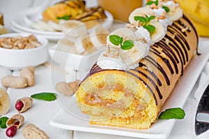 Biscuit or sponge cake roll with peanut butter cream, bananas and chocolate glaze on a white wooden background. Horizontal