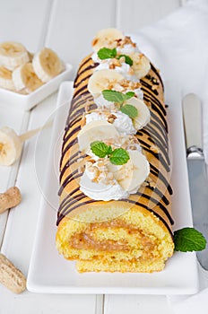 Biscuit or sponge cake roll with peanut butter cream, bananas and chocolate glaze on a white wooden background. Vertical