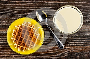 Biscuit waffle in saucer, spoon, transparent bowl with condensed milk on table. Top view