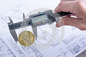 Bitcoin coin is clamped in a digital vernier caliper against a blurry background of drawings