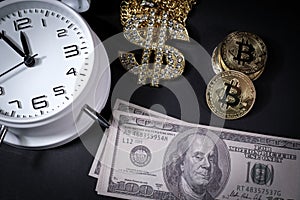 Two Bitcoin coins lie next to a stack of $100 bills, a white alarm clock and a diamond-studded dollar chain pendant
