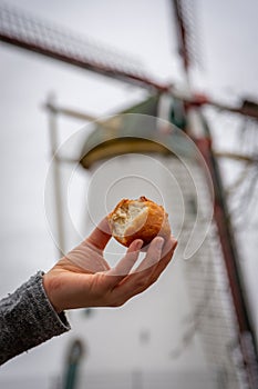 Bite out of typical Dutch wintertime deep fried pastry called `oliebol` held in front of traditional windmill